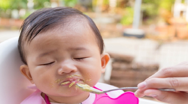 Baby with closed eyes reluctantly tasting food from a spoon.
