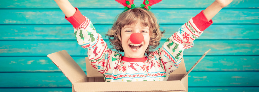 Child in festive reindeer attire with a red nose, smiling joyfully inside a cardboard box.