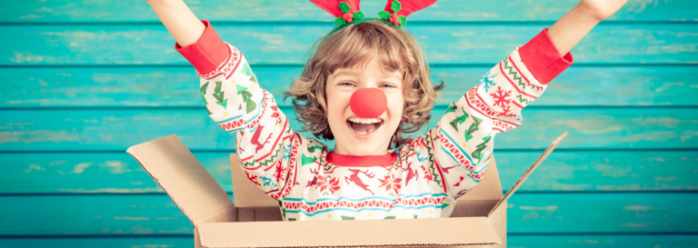 Child in festive reindeer attire with a red nose, smiling joyfully inside a cardboard box.