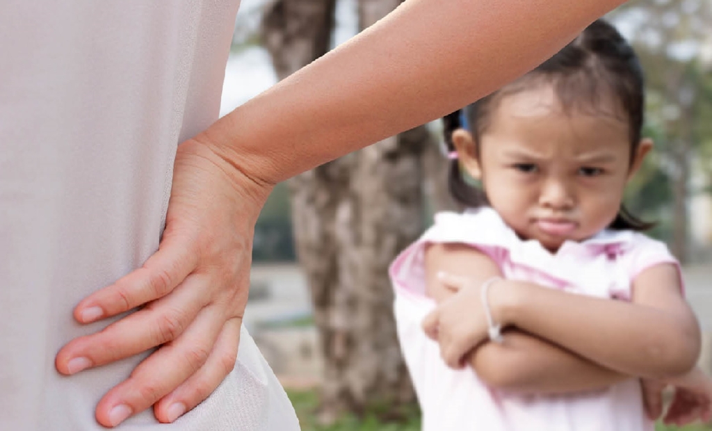 Little girl pouting with crossed arms behind an adult’s hand on hip.