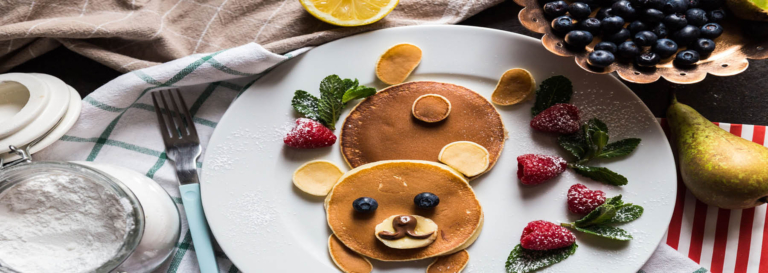 Bear-shaped pancake with fruit and chocolate details, garnished with raspberries and mint leaves.