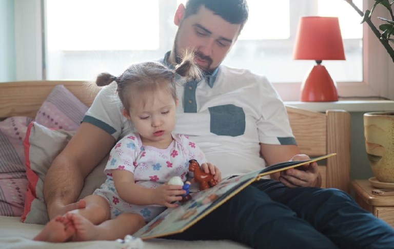 Father reading a book while daughter plays with toys beside him.