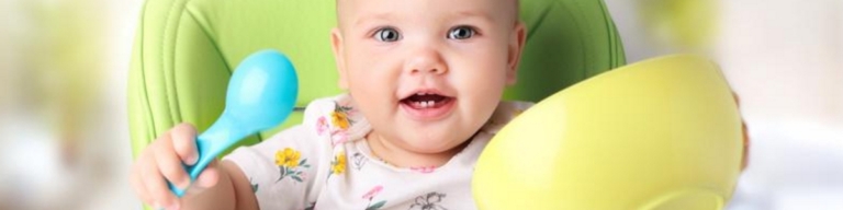 Smiling baby holding a yellow bowl and blue spoon in a high chair.