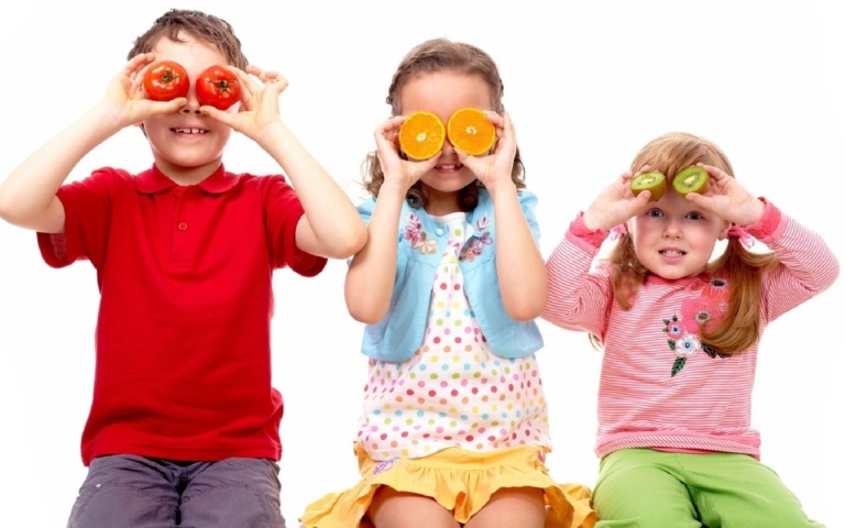 Three kids holding fruits as playful pretend glasses.