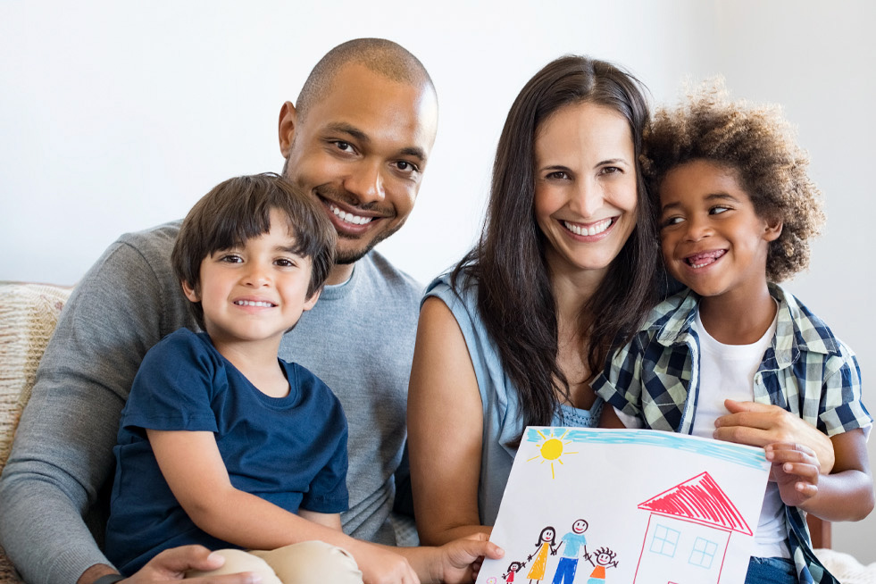 A happy family of four smiling together, with the children holding a colorful drawing.